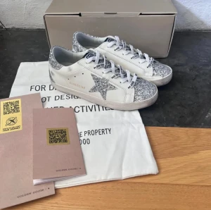 Golden Goose sneakers med glitter - Snygga vita sneakers från Golden Goose med silverglittriga detaljer på tå, häl och stjärna på sidan. Skorna har snörning och platt sula, perfekt för dig som gillar att sticka ut med lite extra bling. Märkeslogga på tungan och sidan.