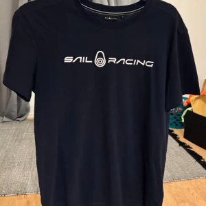 Mörkblå Sail Racing t-shirt i bomull - Snygg mörkblå t-shirt från Sail Racing med vit logga tryckt på bröstet. Klassisk passform och rund halsringning. Tillverkad i mjuk bomull, perfekt för en clean och sportig look. Helt ny 1 gång använd och hälften av priset än att köpa den från affären.