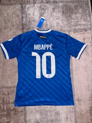 Blå Real Madrid Mbappé tröja Adidas L - Snygg blå Real Madrid fotbollströja från Adidas med Mbappé och nummer 10 på ryggen. Tröjan har vita detaljer, klassiska Adidas-ränder på axlarna, klubbmärke på bröstet och Emirates Fly Better som sponsor. Tillverkad i lätt polyester med korta ärmar.