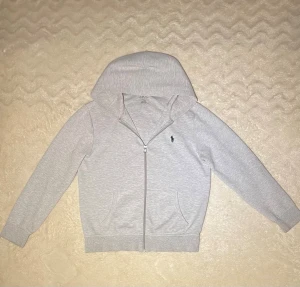 Grå hoodie från Ralph Lauren - Snygg ljusgrå hoodie från Ralph Lauren med dragkedja och huva. Tröjan har långa ärmar, två fickor fram och den klassiska Polo-loggan broderad på bröstet. Perfekt för chill dagar och enkel att matcha med jeans eller joggers.