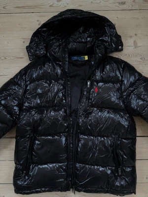 Svart pufferjacka från Polo Ralph Lauren - Säljer en svart, glansig pufferjacka från Polo Ralph Lauren med huva och röda broderade loggan på bröstet. Jackan har dragkedjor på fickorna och framtill, samt quiltad design som ger en riktigt fet vinterlook.
