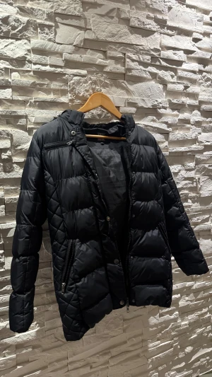 Svart pufferjacka från Vinson med fuskpäls - Svart pufferjacka från Vinson med snygg quiltad design, dragkedjor framtill och huva med brun fuskpäls. Jackan har långa ärmar och en klassisk pufferlook som håller dig varm under kalla dagar. Man kan även ta bort luvan om man inte vill ha med den! 