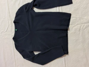 Mörkblå stickad tröja Benetton - Mörkblå stickad tröja från United Colors of Benetton med v-ringad hals och långärmad passform. Diskret broderad logga i blått på nederkanten. Tröjan har ribbade muddar vid ärmslut och nederkant, perfekt för dig som gillar enkel och stilren design.