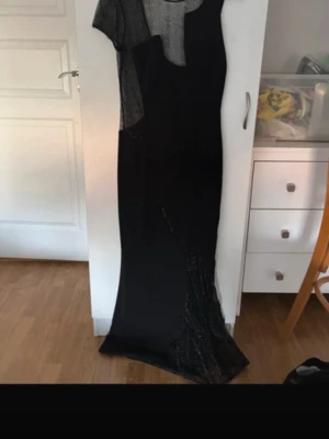 Svart långklänning med meshdetaljer - Elegant svart långklänning med transparenta meshpartier upptill och längs benet. Klänningen har en figurnära passform och är ärmlös med rund halsringning. Perfekt för en snygg och stilren look.