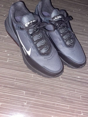 Nike grå och svarta sneakers - Snygga grå och svarta Nike sneakers med vit swoosh på sidan och coolt mönster på plösen. Skorna har snörning och är tillverkade i mesh och syntetmaterial för en sportig look. Perfekta för dig som gillar streetwear och vill ha bekväma skor till vardagen. Oanvända skor.