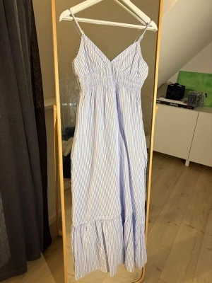 Blåvit långklänning med smala axelband - Säljer en somrig blåvit långklänning med smala axelband och v-ringning. Klänningen har ett smockat liv, tunna vertikala ränder och volang längst ner. Perfekt för varma dagar och enkel att styla med smycken eller sandaler.