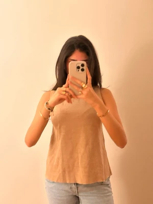 Beige linnetopp i mjukt material - Snygg beige linnetopp med ärmlös design och rund halsringning. Toppen har en enkel och stilren look som passar perfekt till jeans eller kjol. Materialet ser mjukt och bekvämt ut, perfekt för varmare dagar.