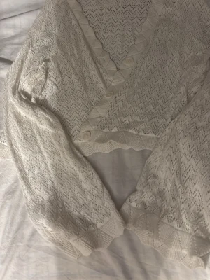 Vit spetsblus med knappar - Söt vit kofta i spets med v-ringning och dekorativa knappar framtill. Koftan har ett fint mönster, vågiga kanter och långa ärmar. Perfekt för dig som gillar romantisk och vintage-inspirerad stil.