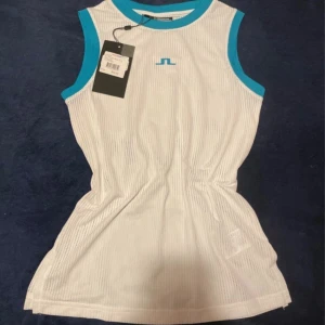 Vit mesh linnetopp från J.Lindeberg - Sportig vit mesh linnetopp från J.Lindeberg med turkosa kanter och logga framtill. Toppen är ärmlös och har en luftig, perforerad design som passar perfekt för träning eller tennis. Materialet är syntet och ger en fräsch känsla under aktiva dagar.