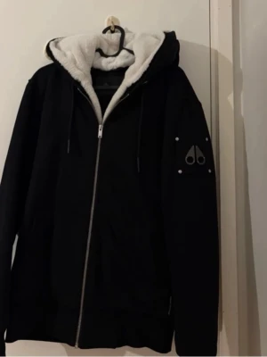 Svart Tröja från Moose Knuckles - Snygg svart hoodie från Moose Knuckles med dragkedja och vit fluffig foder på insidan av huvan. Märkets logga sitter på ärmen och luvan har dragsko. Perfekt för dig som vill ha en varm och stilren hoodie med exklusiv känsla.
