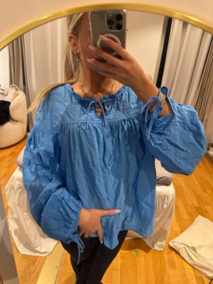 Blå voluminös blus med knytband - Snygg blå blus med voluminösa, långa ärmar och knytband vid halsringningen och ärmsluten. Blusen har en lös passform och är tillverkad i ett lätt, skrynkligt tyg som ger en avslappnad vibe. Perfekt att styla med jeans eller kjol.