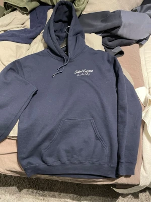 Mörkblå Saint Tropez hoodie - Mörkblå hoodie från Saint Tropez Yacht Club med broderad logga på bröstet och stor färgglad print av en båt på ryggen. Storlek S men passar XS med. Nypris 800 knappt använd.