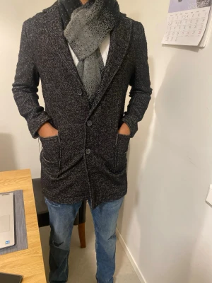 Mörkgrå ullkappa med knappar - Stilren mörkgrå kappa i ullblandning med klassisk krage och stora framfickor. Kappan har knäppning med tre knappar framtill och en rak, något längre passform. Perfekt för kyliga dagar och ger en snygg, tidlös look.