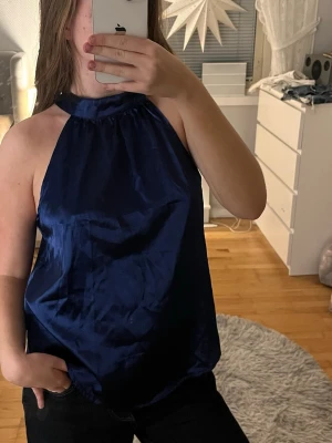 Mörkblå halterneck topp i satin - Snygg mörkblå halterneck topp i glansigt satinliknande material. Toppen är ärmlös med hög hals och lös passform, perfekt för en trendig och stilren look.