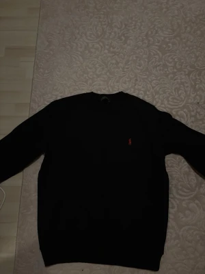 Svart stickad tröja från Ralph Lauren - Klassisk svart stickad tröja från Ralph Lauren med den ikoniska röda logotypen broderad på bröstet. Tröjan har rund halsringning och långa ärmar, perfekt för en stilren och enkel look.