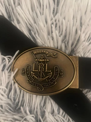 Svart läderbälte från Ralph Lauren - Snyggt svart bälte i läder från Ralph Lauren med en oval, antikguldfärgad metallspänne. Spännet har ett detaljerat ankare och flagga med texten 'Ralph Lauren Est. 1967'. Perfekt accessoar för att lyfta din outfit.
