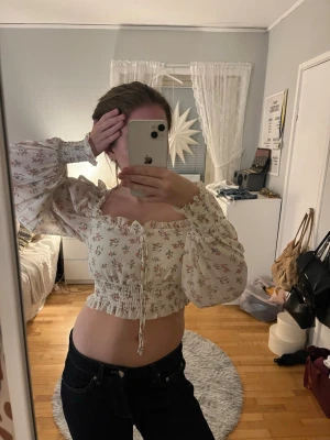 Blommig croppad blus med puffärmar - Supersöt blommig croppad blus i vitt med rosa och gröna detaljer. Toppen har långa puffärmar, smockad midja och knyt framtill. Perfekt för dig som gillar romantisk och trendig stil.