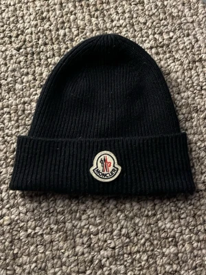 Svart Moncler mössa i ull - Säljer en svart ribbstickad mössa från Moncler med klassisk logga framtill. Mössan är tillverkad i mjuk ull och har en vikt kant för extra stilpoäng. Perfekt accessoar för kyliga dagar och snygg till alla outfits.