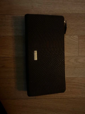 Svart plånbok By Bianca Ingrosso - Säljer en svart plånbok från By Bianca Ingrosso med reptilmönstrad yta och guldfärgad logga framtill. Plånboken har dragkedja runtom och är tillverkad i ett material som liknar skinn. Perfekt storlek för att få plats med allt du behöver.