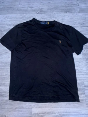 Svart t-shirt från Polo Ralph Lauren - Klassisk svart t-shirt från Polo Ralph Lauren i custom slim fit. T-shirten har korta ärmar och den ikoniska broderade polospelaren på bröstet. Tillverkad i mjuk bomull som känns skön mot huden. Perfekt till jeans eller shorts för en clean look. Storleken på tröjan är L men den passar M med.