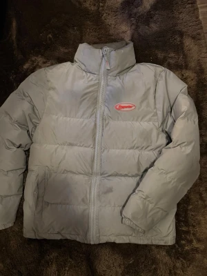 Grå pufferjacka från Trapstar - Säljer en grå pufferjacka från Trapstar med stor röd logga på ryggen och mindre logga på bröstet. Jackan har hög krage, dragkedja framtill och två sidofickor. Perfekt för kalla dagar och riktigt streetwear-vibe.