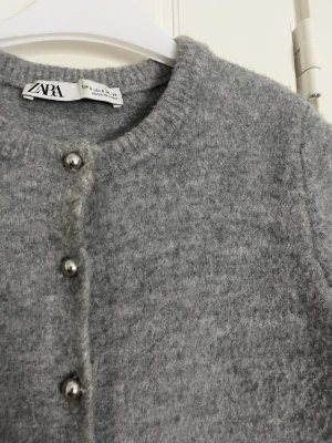 Grå kortärmad kofta från Zara - Mysig grå kofta från Zara med korta ärmar och rund hals. Koftan är stickad i mjukt material och har silvriga klotknappar framtill. Säng till i fall ni vill ha fler bilder,mått eller vill se hur den sitter på💗