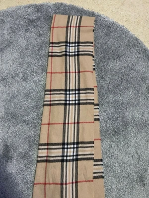 Beige rutig halsduk med fransar - Snygg beige halsduk med klassiskt rutigt mönster i svart, vitt och rött. Halsduken har fransar i ändarna och är tillverkad i ett mjukt material som känns skönt mot huden. Perfekt accessoar för att lyfta din outfit.