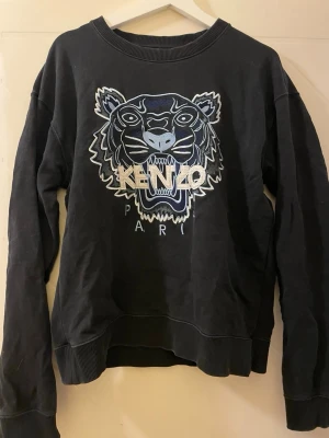 Svart sweatshirt från Kenzo med tiger - Svart sweatshirt från Kenzo med ikonisk broderad tiger och texten 'KENZO PARIS' framtill. Tröjan har ribbade muddar vid ärmslut och nederkant samt rund halsringning. Tillverkad i mjuk bomull för skön känsla och stilren look. Storlek. Lx men sitter som  M - L