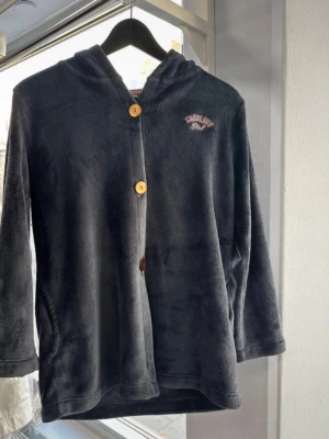 Mörkgrå fleecekofta med huva - Mysig mörkgrå kofta från Christine Bernadotte i mjuk fleece. Har huva, stora träknappar och broderad text 'Fabulous Girl' på bröstet. Perfekt för kyliga dagar och enkel att soppa över en t-shirt. Långärmad och med fickor på sidorna.