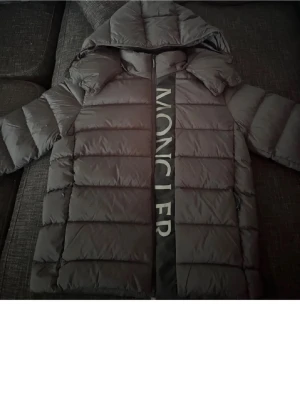 Svart dunjacka från Moncler - Snygg svart dunjacka från Moncler med stor logga längs dragkedjan. Jackan har en puffig puffer-look, huva och långa ärmar. Perfekt för kalla dagar och riktigt trendig stil.