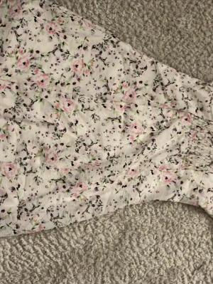 Blommig kortklänning med puffärm - Söt kortklänning med puffärmar i vitt tyg med rosa och grönt blommönster. Klänningen har smock i midjan och en lätt utsvängd kjol. Perfekt för sommarens alla tillfällen!