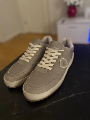 Grå sneakers från Philippe Model - Helt nya Philippe modells. Snygga grå sneakers från Philippe Model med vita snören och detaljer i mocka. Skorna har en klassisk låg siluett, perforerad tå och vit sula. Perfekta för dig som gillar stilrena och tidlösa sneakers.