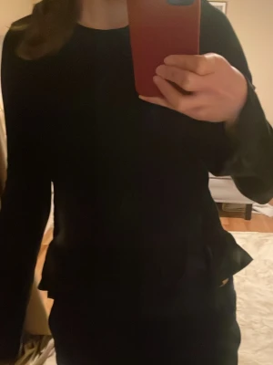 Svart långärmad topp med volang - Svart långärmad topp med volangdetalj nedtill. Enkel och stilren design med rund halsringning. Perfekt att matcha med jeans eller kjol för en snygg look.Gina Young S170