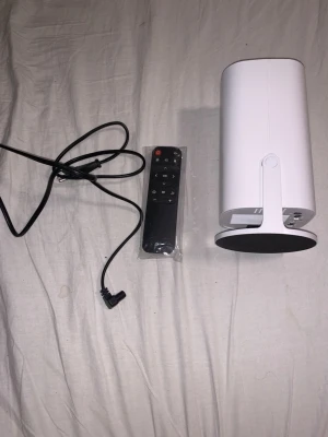 Vit smart projektor med fjärrkontroll - Säljer en vit smart projektor med rund fot, tillhörande svart fjärrkontroll och strömkabel. Projektorn har en modern, cylinderformad design och passar perfekt för filmkvällar eller gaming. Enkel att styra med fjärrkontrollen och smidig att placera tack vare det kompakta formatet.