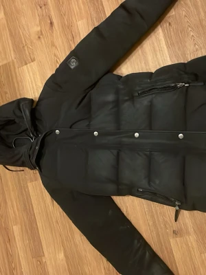 Svart dunjacka med huva och dragkedjor - Snygg svart dunjacka från Save The Duck med huva och silverfärgade dragkedjor på fickorna. Jackan har quiltad design, långa ärmar och tryckknappar framtill. Perfekt för kalla dagar och enkel att matcha med allt. Den har används några gånger men går att tvätta! Pris kan diskuteras.