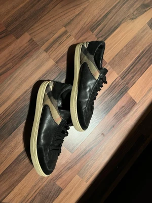 Burberry skor - Snygga svarta sneakers med beige och röd rand längs sidan. Skorna har klassisk låg profil, svarta skosnören och vit sula. Materialet är en mix av skinn och textil, vilket ger en stilren och modern look. PRIS KAN DISKUTERAS.