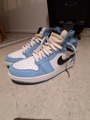 Nike Air Jordan 1  - Snygga Nike Air Jordan 1 sneakers i blått och vitt med svarta detaljer. Klassisk hög modell med snörning och rund tå. Ovandelen är i skinn och har perforerade partier för extra ventilation. Perfekta för dig som vill ha en ikonisk och stilren sneaker.