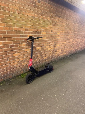 Kukirin g2 - Säljer en svart elscooter med robusta däck och en röd bandana knuten runt styret. Scootern har bred fotplatta med halkskydd och stöd. Perfekt för att ta sig runt i stan på ett smidigt sätt. 