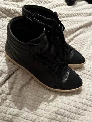 Svarta höga sneakers med snörning från Ecco - Snygga svarta sneakers med högt skaft, snörning och dragkedja på sidan. Skorna har vit sula och är tillverkade i skinnliknande material. Perfekta för dig som gillar en stilren och urban look.