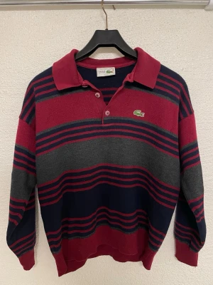 Randig tröja från Lacoste i ullmix - Snygg retroinspirerad tröja från Lacoste med breda ränder i vinrött, mörkblått och grått. Tröjan har klassisk krage, knappslå och den ikoniska krokodilloggan på bröstet. Tillverkad i mjuk ullmix och har ribbade muddar vid ärmslut och nederkant.