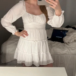 Vit kort klänning med volang - Supersöt vit kort klänning med volangkjol och långa puffärmar. Klänningen har en markerad midja och lätt rynkade detaljer upptill. Perfekt för sommarens alla tillfällen och ger en romantisk vibe.