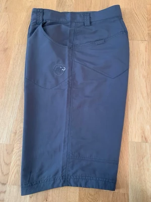 Mörkgrå Mammut shorts  - Säljer ett par mörkgrå shorts från Mammut i storlek 38.De har normal passform, praktiska fickor fram och bak, bälteshällor och dragkedja med dubbla knappar. Materialet är slitstark syntet, perfekt för utomhusaktiviteter.