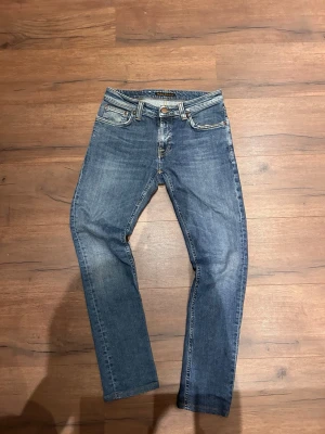 Blå jeans från Nudie Jeans - Säljer ett par klassiska blå jeans från Nudie Jeans med slim passform. Jeansen har fem fickor, gylf med dragkedja och Nudie-logga på bakfickan. Tillverkade i slitstark bomull med snygg tvättad look. Är en slitning i fickan men inget som syns vid användning och dem är slim på mig men jag är dock riktigt smal och kanske sitter tajtare på dig