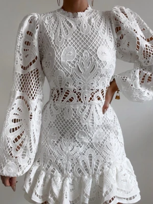 Vit spetsklänning med volang  - Superfin vit kort klänning med broderade spetsdetaljer och volang nertill. Klänningen har långärmade puffärmar och vackert mönster i spetsen. Hög halsringning och markerad midja ger en snygg siluett. Perfekt för sommarens alla tillfällen.