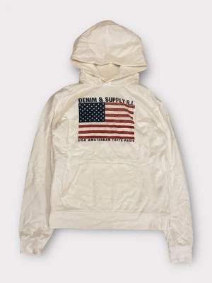 Vit hoodie Denim & Supply R.L. Ralph Lauren  - Vit hoodie från Denim & Supply R.L. med stor amerikansk flagga och text på bröstet. Trycket inkluderar städer som USA, Amsterdam, Tokyo och Paris. Hoodien har huva och magficka, samt ribbade muddar vid ärmslut och nederkant. Perfekt för en avslappnad streetwear-look.
