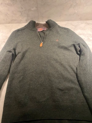 Grön halvzip tröja från Morris - Mörkgrön stickad tröja från Morris med halv dragkedja och liten broderad logga på bröstet. Tröjan har hög krage och ribbade muddar vid ärmslut och nederkant. Perfekt för lager på lager och snygg till jeans.
