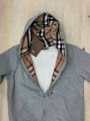 Grå hoodie med Burberry-mönstrad huva - Snygg grå zip-hoodie från Burberry med klassiskt rutigt foder i huvan. Hoodien har dragsko, två fickor framtill och ribbade muddar. Perfekt för dig som gillar stilrena plagg med en twist. Materialet är mjukt och bekvämt, troligen bomullsblandning.