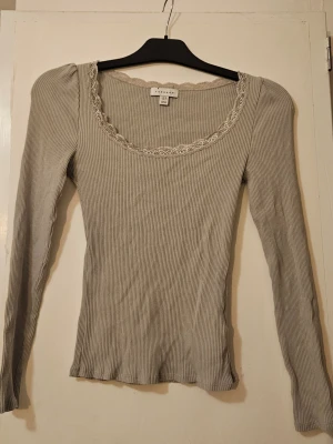 Ljusgrå ribbad topp  - Supersöt ljusgrå ribbad topp med lång ärm och spetsdetalj runt den breda, rundade urringningen. Perfekt att styla med jeans eller kjol för en trendig look. Materialet är mjukt och stretchigt för skön passform.