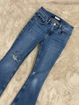 Blå bootcut jeans med slitningar - Snygga blå bootcut jeans med slitna detaljer på låret och knät. Klassisk femficksmodell med knapp och dragkedja framtill. Jeansen har en lätt utsvängd passform nedtill och är tillverkade i denim för en avslappnad look. Den går inte som en 38 egentligen, jag är 34,36 i jeans och passar mig perfekt. 
