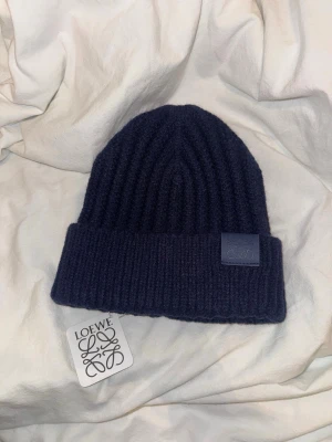 Mörkblå ribbstickad mössa Loewe - Snygg mörkblå ribbstickad mössa från Loewe i lyxig mix av kashmir och ull. Klassisk uppvikt kant med Loewe-logga på en patch framtill. Perfekt accessoar för kalla dagar och riktigt stilren look.